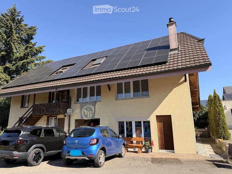 Two-story house with solar panels on the roof, a balcony, and a wooden bench near the entrance. Two cars are parked in front.