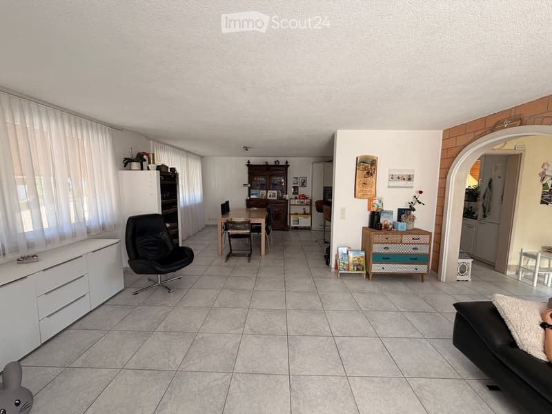 A spacious living and dining area with tiled flooring, large windows with white curtains, a black leather sofa, a dining table with chairs, a white cabinet, a refrigerator, and various decorative items including picture frames and plants.