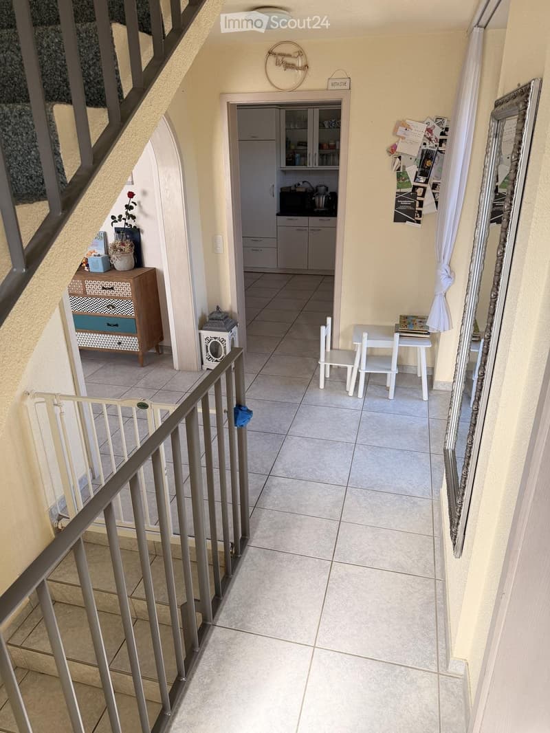 Balcony with tiled floor, small table and chairs, cabinet with decorative items, and a kitchen area visible in the background.