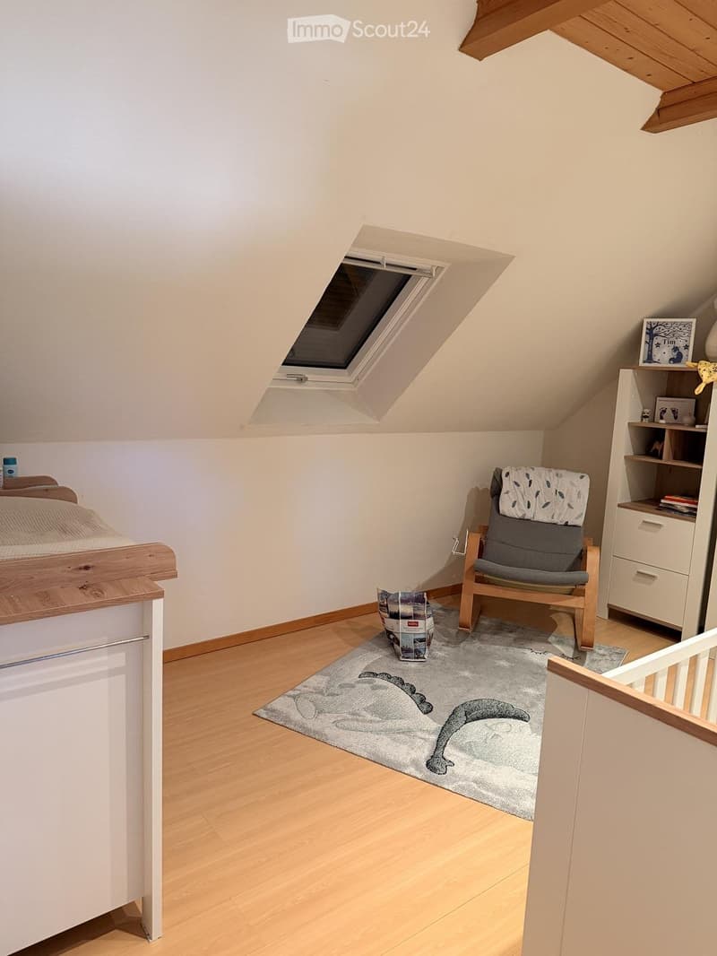 Cozy attic bedroom with wooden floor, slanted ceiling, skylight window, bed, armchair, shelf, and rug with dinosaur print.