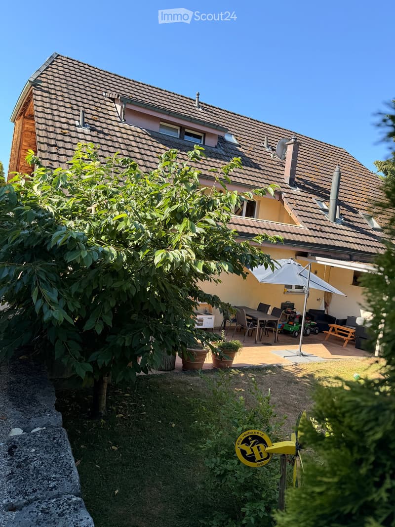 Garden area with a patio, outdoor furniture, umbrella, and a small tree. The house has a tiled roof and multiple windows.