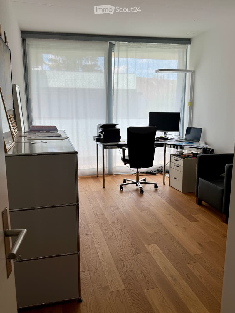 Office space with wooden flooring, large window with blinds, desk with chair, laptop, printer, and a sofa in the corner.