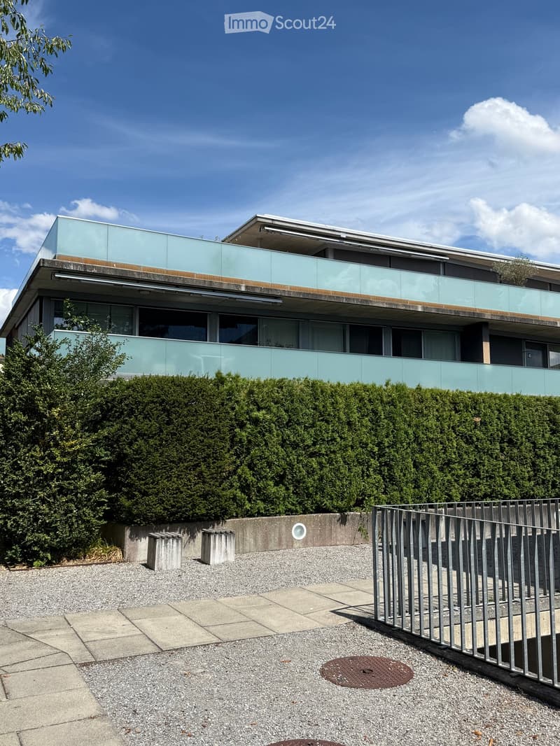 Modern two-story building with large glass windows and balconies, surrounded by greenery and a gravel area.