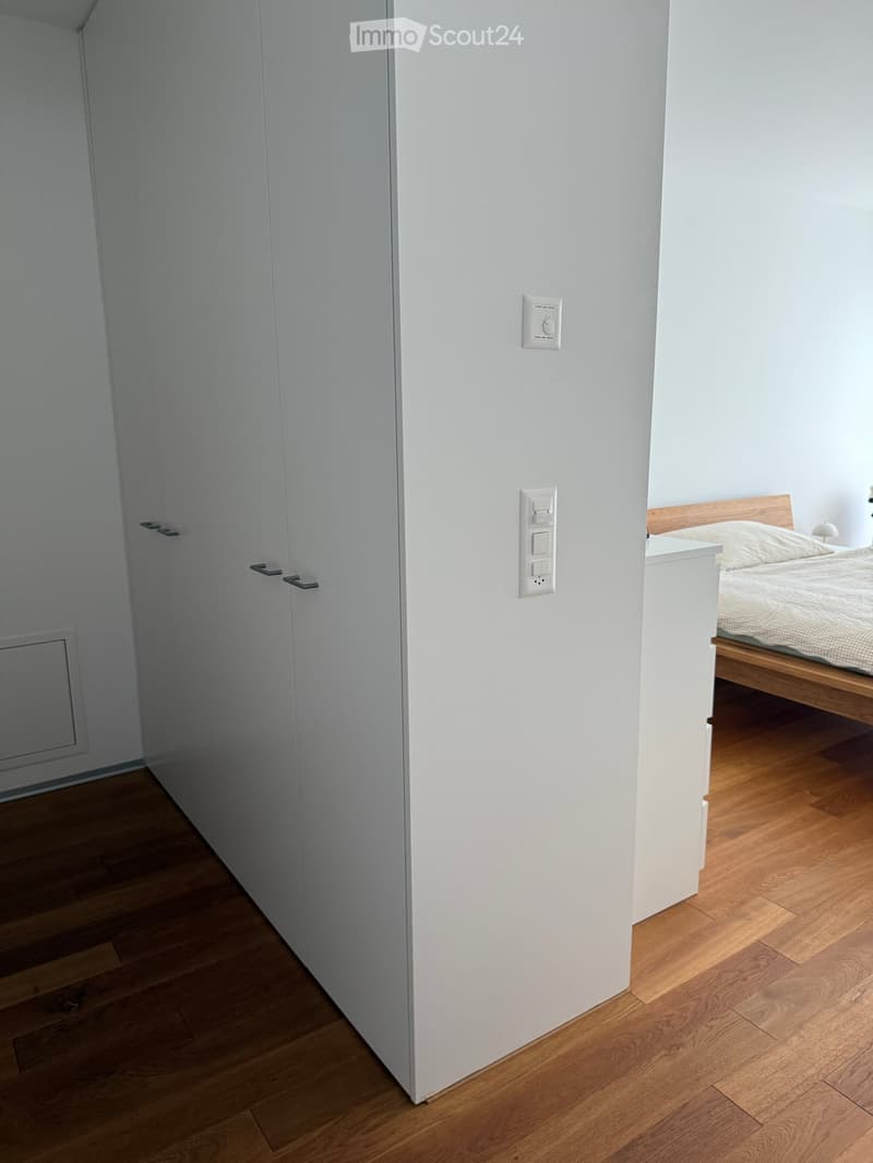 Modern bedroom with light wooden flooring, white walls, a bed with a wooden frame, and a built-in wardrobe with sleek handles.