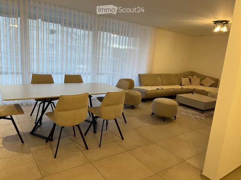 Modern living room with a sofa, coffee table, ottoman, and dining area with a table and chairs. Large windows with sheer curtains provide natural light.