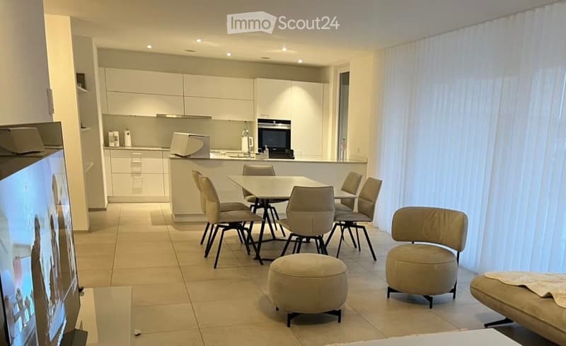 Modern, open-plan living and dining area with a sofa, ottoman, dining table, chairs, and a fully equipped kitchen with cabinets, oven, and fridge.
