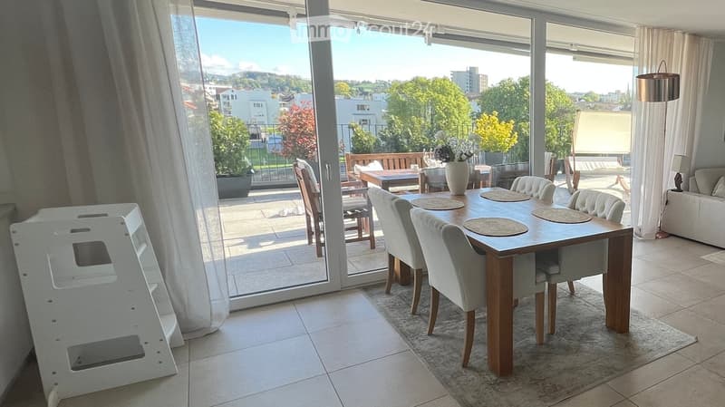 Bright dining area with a wooden dining table, six chairs, a sofa, and a vase on a side table, leading to a balcony with outdoor seating and a scenic view.