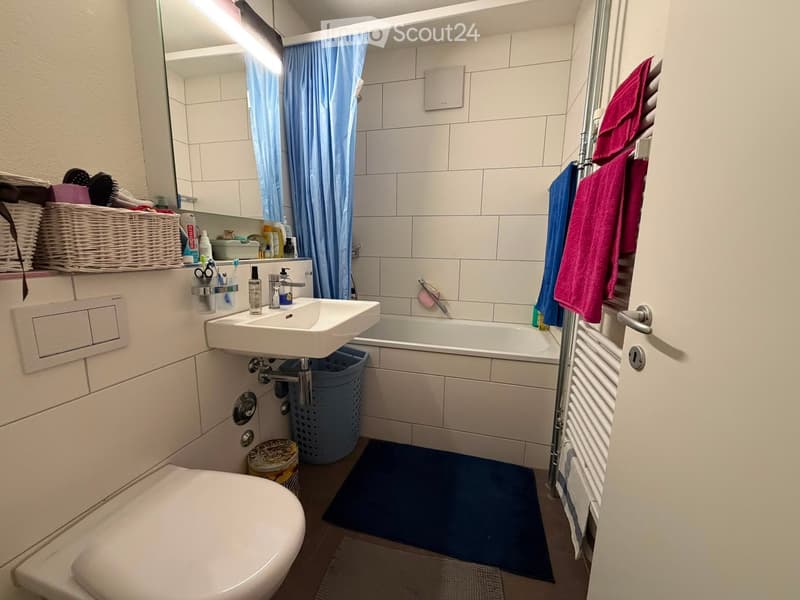 Modern bathroom with white tiles, a sink, toilet, bathtub, mirror, towel rack, and a wicker basket on the counter.