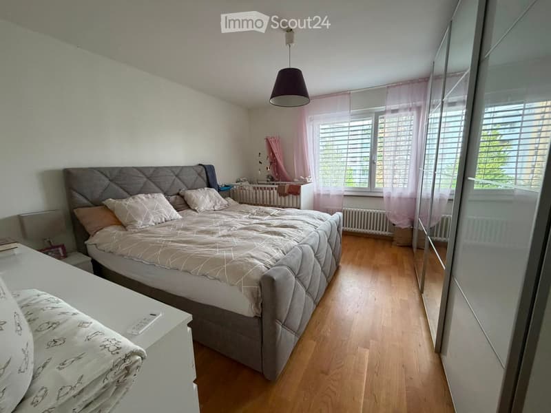 A well-lit bedroom with a large bed, nightstand, mirror, and a small baby crib in the corner. The room features wooden flooring, white walls, and pink curtains on the windows.