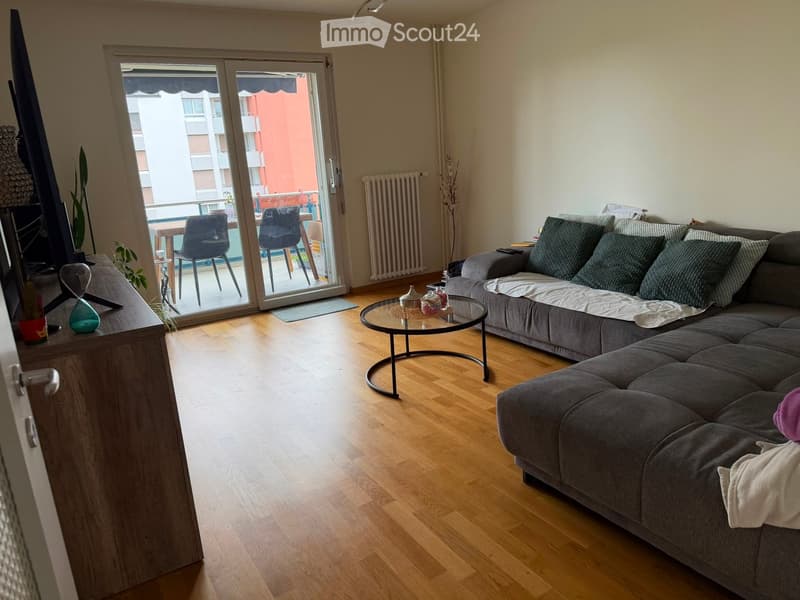 Modern living room with wooden flooring, grey sectional sofa, glass coffee table, and sliding glass door leading to a balcony.
