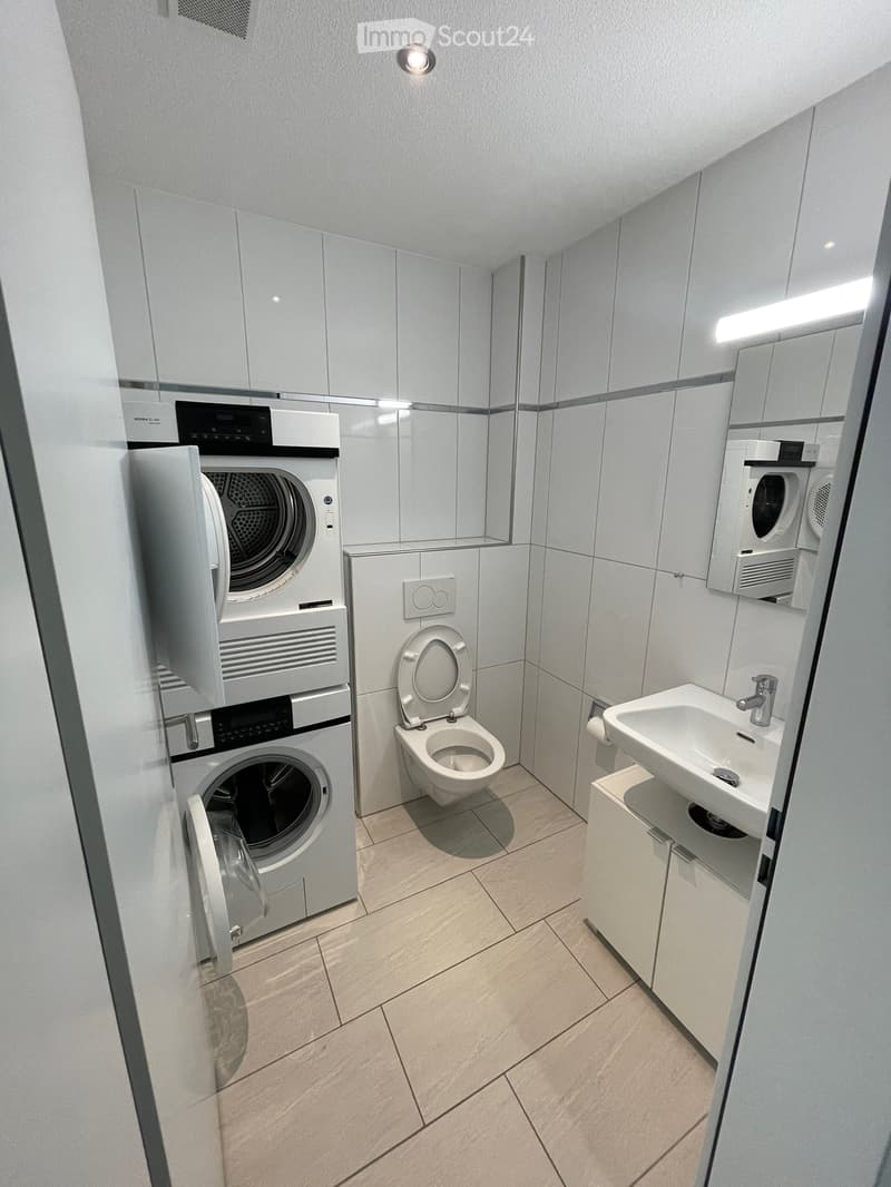 Modern bathroom with a toilet, sink, washing machine, and dryer. Light-colored tiles and cabinets.