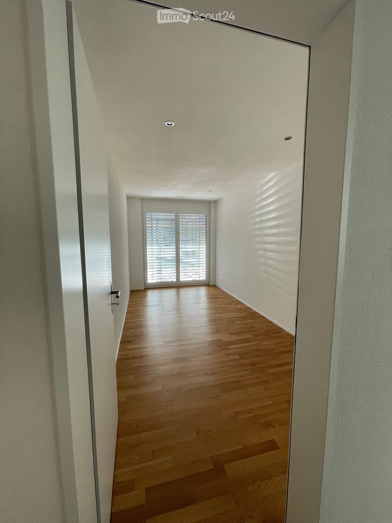 Empty hallway with wooden flooring, white walls, and a large window with blinds at the end.