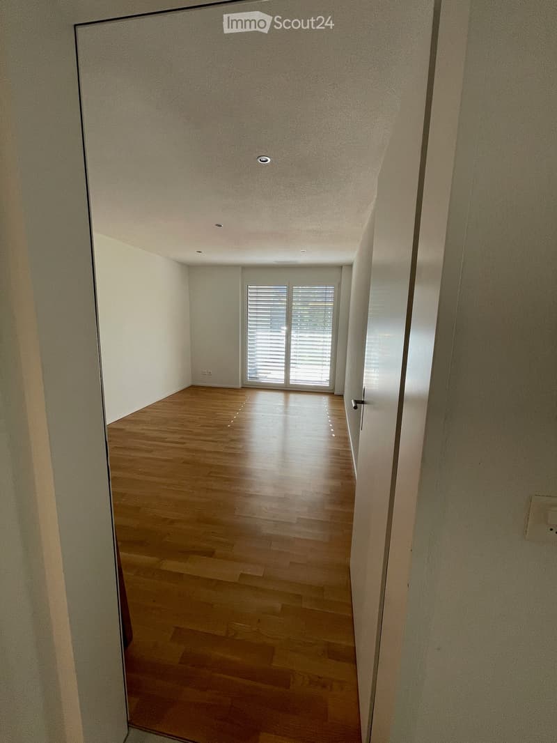 Empty room with wooden flooring, white walls, and a large window with blinds at the far end.