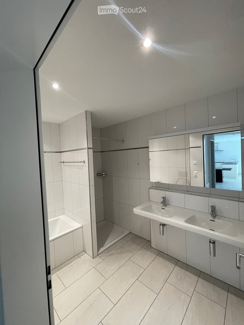 Modern bathroom with a double sink, large mirror, bathtub, and built-in storage. The space features light grey tiles and a sleek, contemporary design.