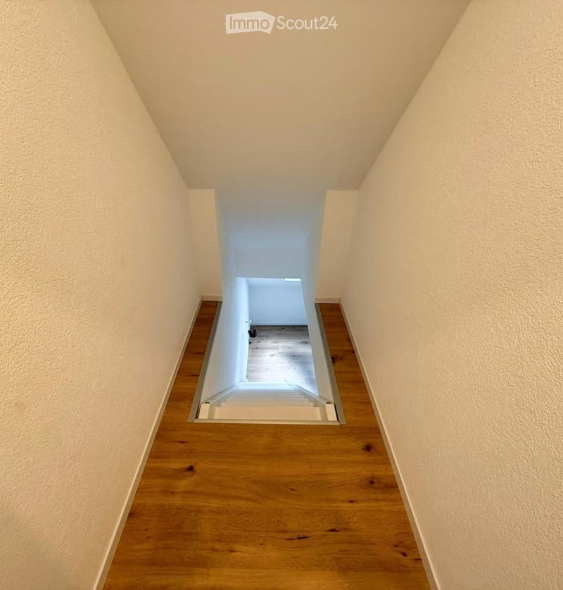 A narrow hallway with wooden flooring and white walls, leading to a bright open space at the end.