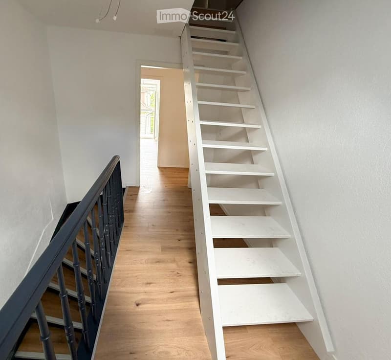 A narrow hallway with wooden flooring and white walls, featuring a staircase with white steps and a dark wooden railing on the left side. A doorway leads to another room at the end of the hallway.