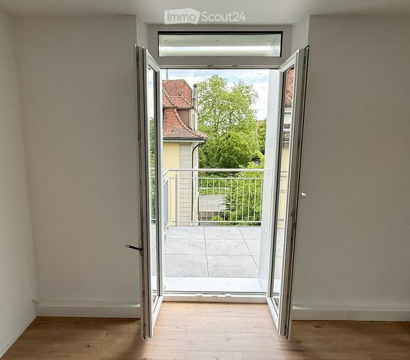 Empty room with wooden flooring and large open sliding doors leading to a balcony with a view of a neighboring building and greenery.