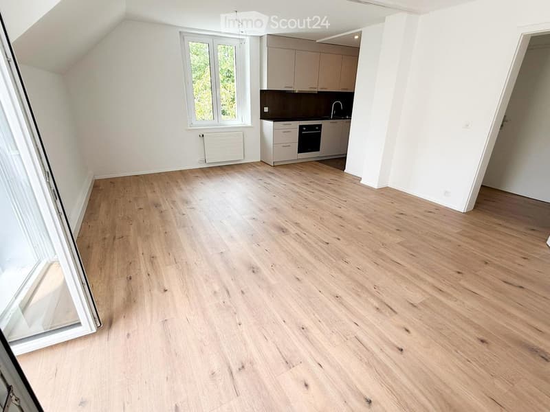 Empty room with light wooden flooring, white walls, and a small kitchen area with cabinets and appliances.
