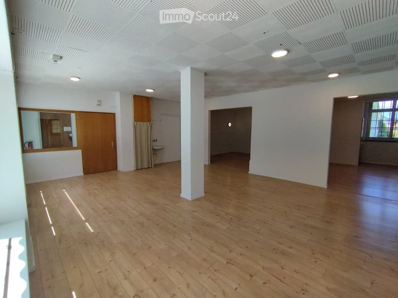 Empty room with wooden flooring, white walls, and a central column. Features include a wooden door, a small sink area, and a window with a curtain.