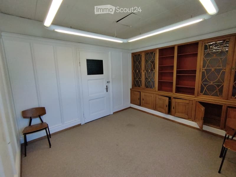 A small, unfurnished room with a wooden cabinet and a single chair, featuring white walls and a carpeted floor.