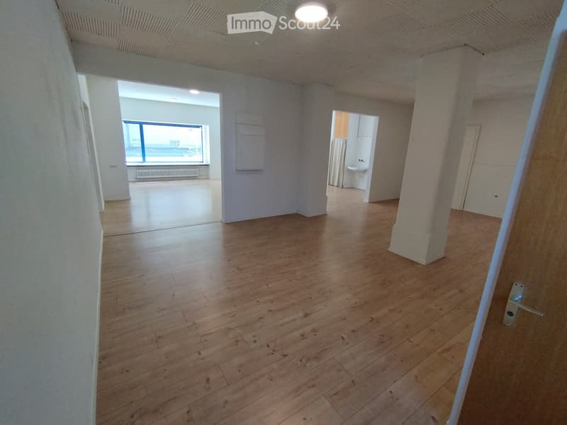 Empty room with wooden flooring, white walls, and a large window with a view. A small adjacent bathroom is visible with a sink and a curtain.