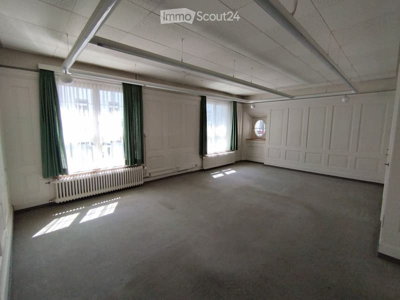Empty room with bare walls, carpeted floor, large windows with green curtains, radiators, and white paneling on one wall.