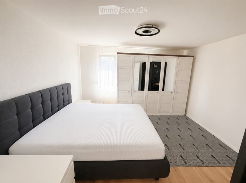 Modern bedroom with a large bed, grey upholstered headboard, white wardrobe with mirrored doors, and a patterned rug on wooden flooring.