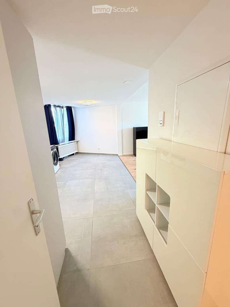 Empty hallway with white walls, tiled floor, and a built-in shelf unit on the right side. A door is visible on the left, and a washing machine is partially visible in the background.