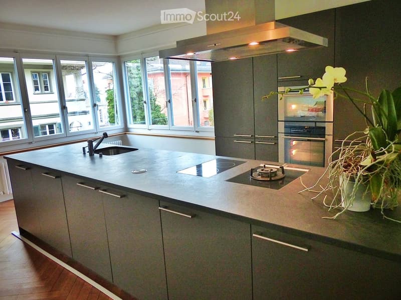 Modern kitchen with grey cabinets, a central island with a sink and stove, and large windows overlooking the street.