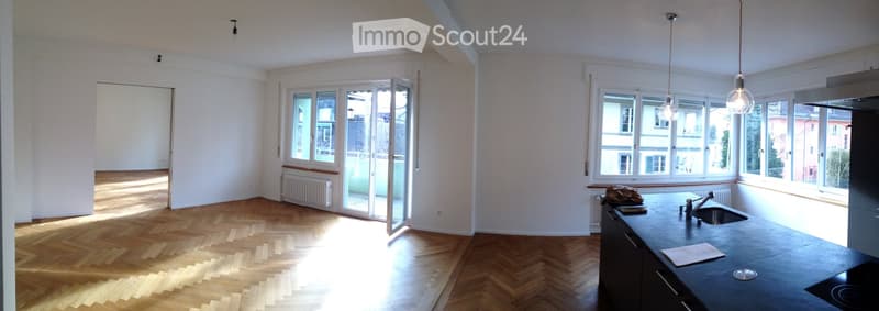 Empty room with wooden flooring, large windows, and a kitchen area with a sink and countertop.
