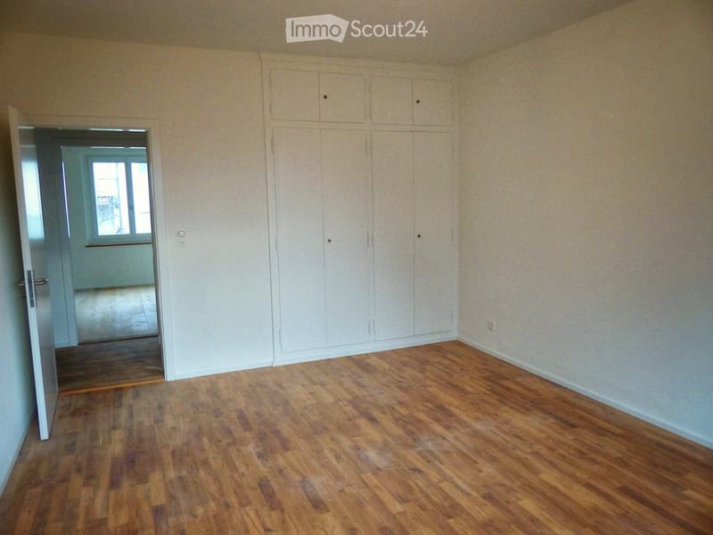 Empty room with wooden flooring, white walls, built-in wardrobes, and an open doorway leading to another room with a window.