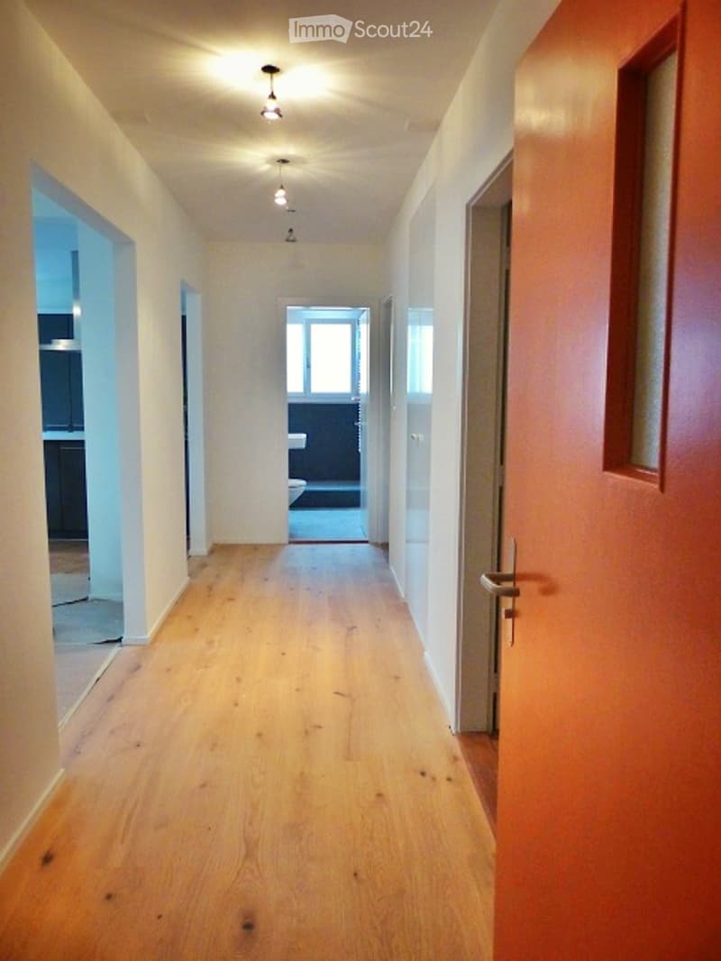 Empty hallway with wooden flooring, white walls, and recessed ceiling lights. Doors lead to adjacent rooms, including a bathroom visible at the end.
