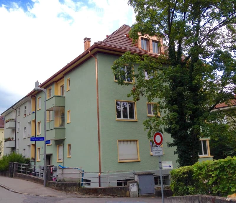 A three-story residential building with green siding and yellow window frames, featuring multiple windows and a small balcony on the second floor.
