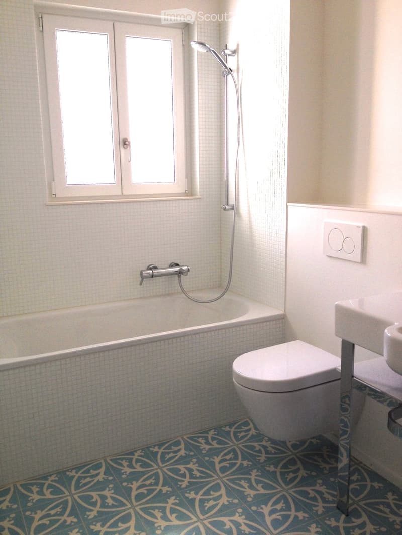 Modern bathroom with white tiled walls, blue and gold patterned floor tiles, bathtub, toilet, sink, and showerhead.