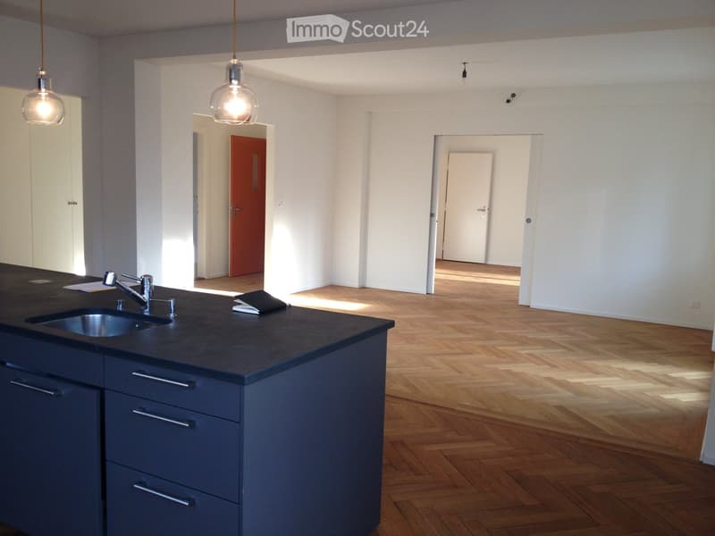 Modern kitchen with dark countertops, stainless steel sink, and dark blue cabinets. Wooden flooring and pendant lights visible.