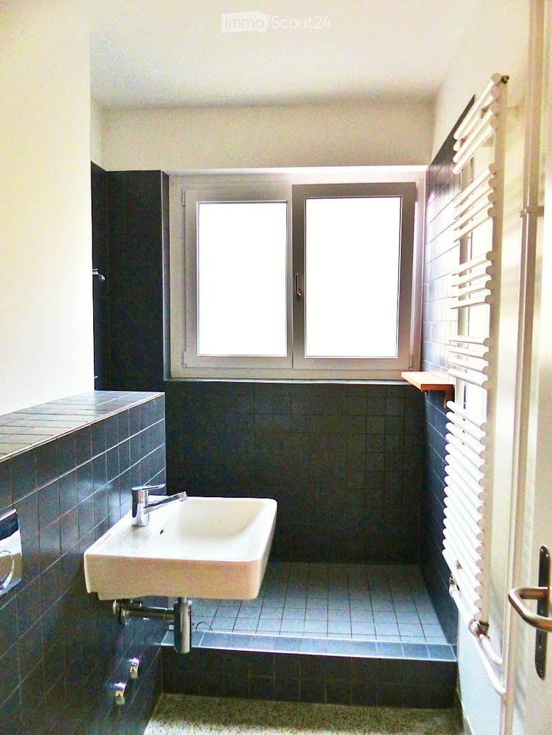 Modern bathroom with a white sink, chrome faucet, dark tiled walls, and a window with blinds.