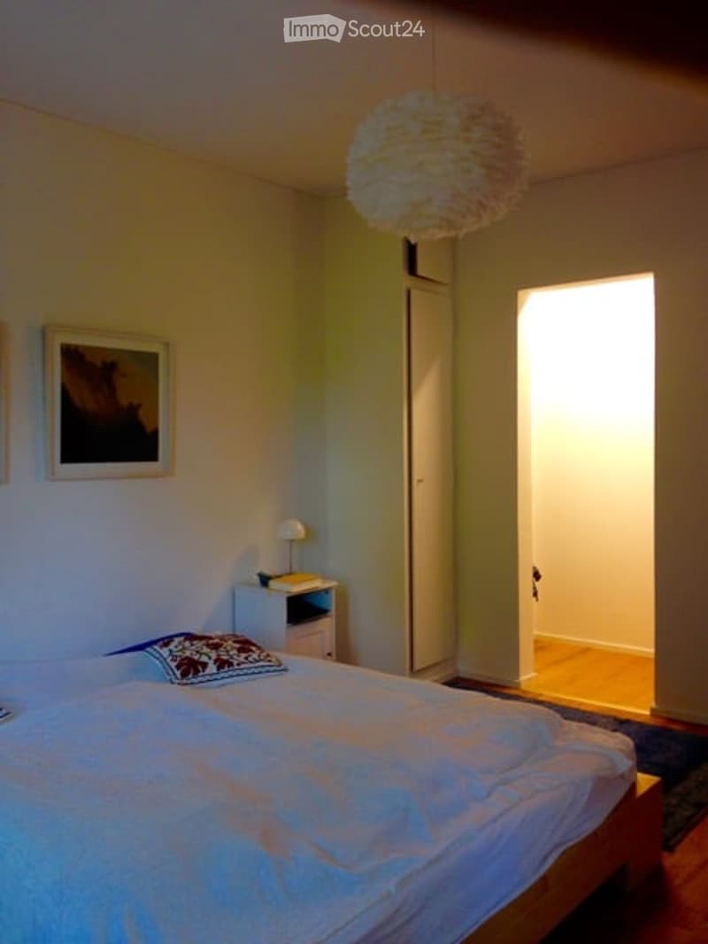 Bedroom with a neatly made bed, nightstand with a lamp, picture frame on the wall, and a hanging decorative light fixture.