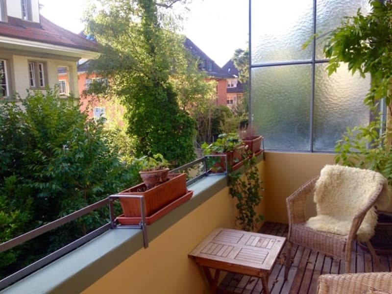 Balcony with wooden floor, wicker chair, small table, and potted plants, overlooking a green area with trees and neighboring buildings.