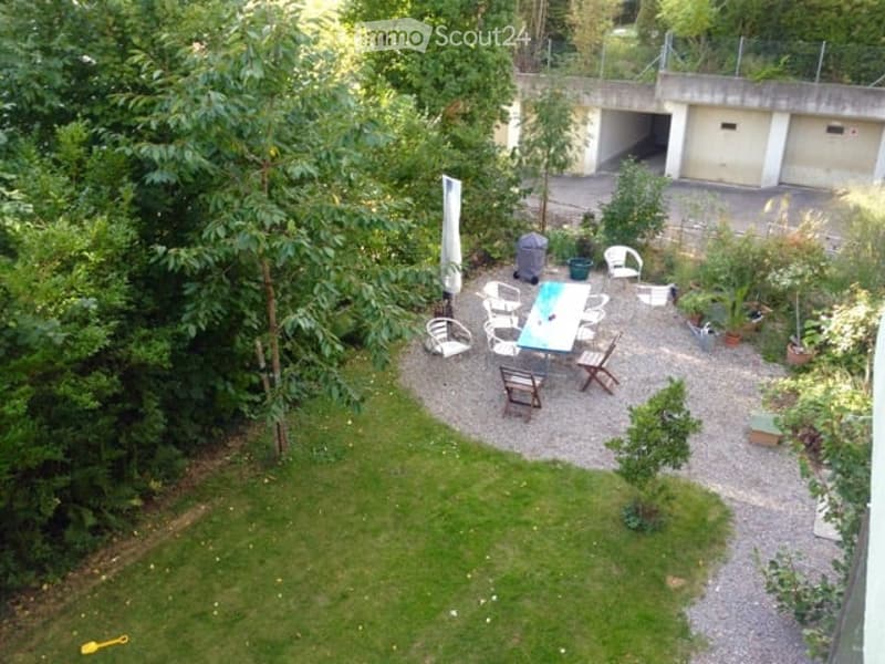Garden area with a table, chairs, and plants, surrounded by trees and a garage in the background.