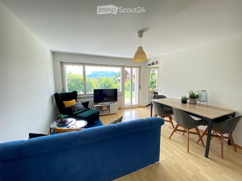 Bright living room with wooden flooring, large windows, and a view of greenery. Features a blue sofa, green armchair, coffee table, dining table with chairs, and a television on a stand.