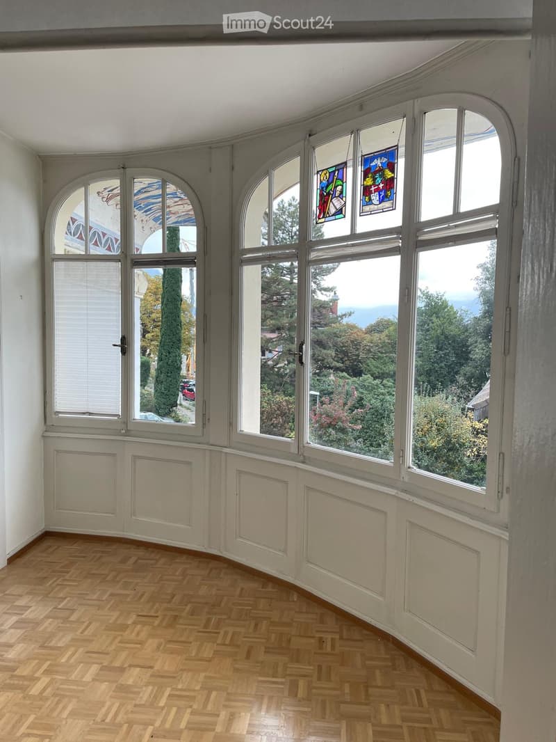 An unfurnished room with large arched windows, wooden herringbone flooring, and white walls. The windows offer a scenic view of trees and a distant landscape.