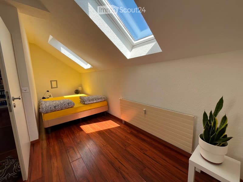 Cozy attic bedroom with wooden flooring, slanted ceiling, skylight windows, a single bed with yellow bedding, a small table with a potted plant, and a radiator.