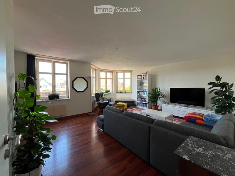Living room with hardwood flooring, large windows, grey sofa, coffee table, television, bookshelf, and plants.