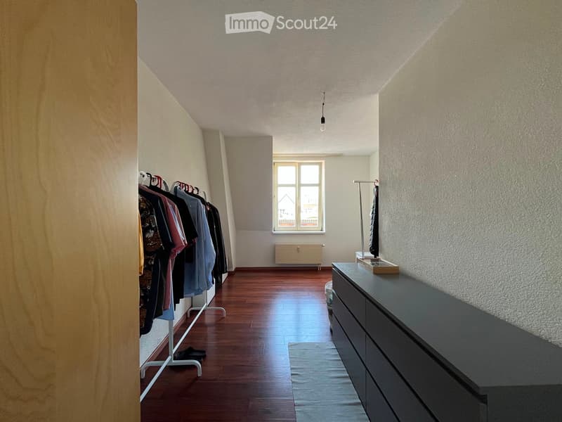 A narrow hallway with wooden flooring, a clothing rack on the left, a dresser on the right, a window at the far end, and a radiator below it.