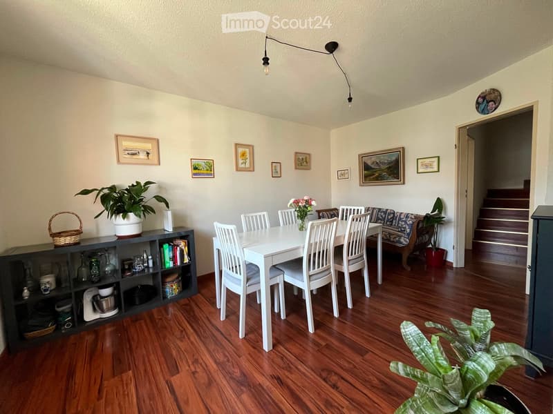 Dining area with a white dining table and chairs, a sofa, a black cabinet with various items, and several potted plants. The room has wooden flooring and several framed pictures on the walls.