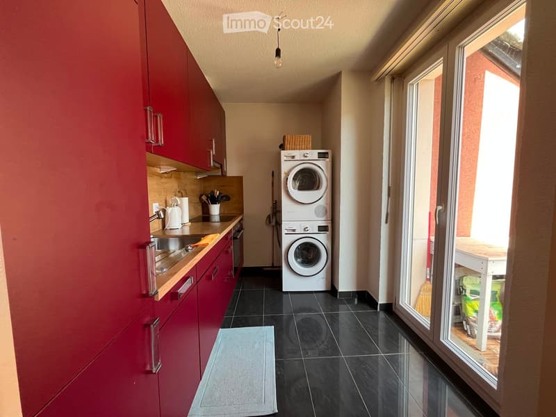 A compact, modern kitchen area with red cabinets, a white washing machine, and a small table near the window.