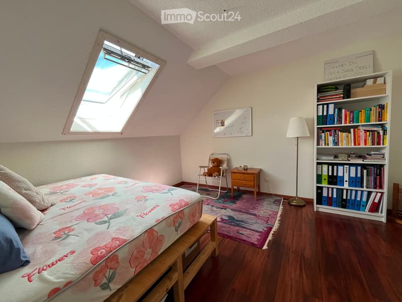 Cozy bedroom with a floral-patterned bed, wooden floor, bookshelf filled with books and binders, small wooden table, floor lamp, and a teddy bear on a chair.