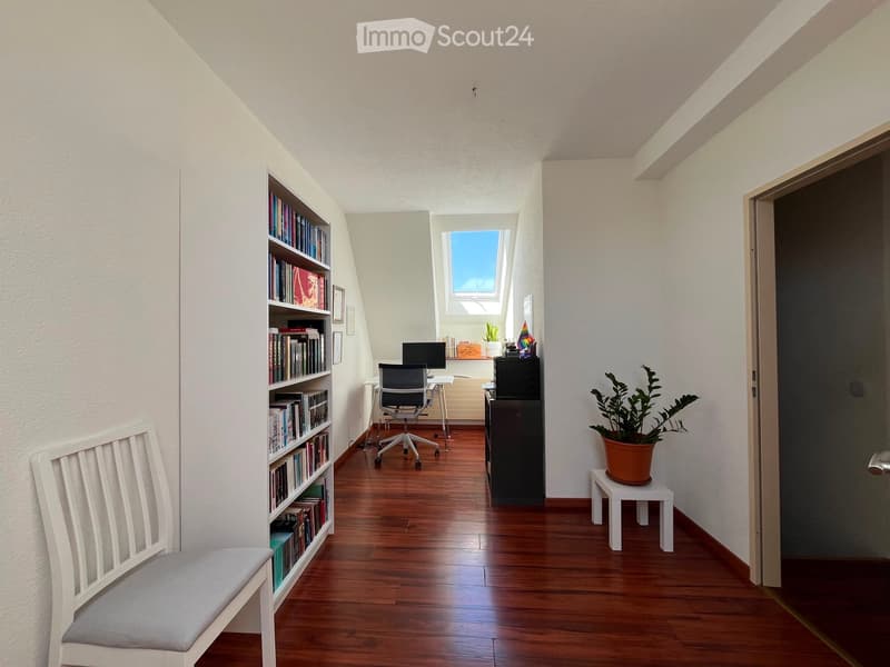 A cozy, well-lit room with wooden flooring, a bookshelf filled with books, a desk with a computer, a chair, a small stool with a potted plant, and a white bench with a cushion.