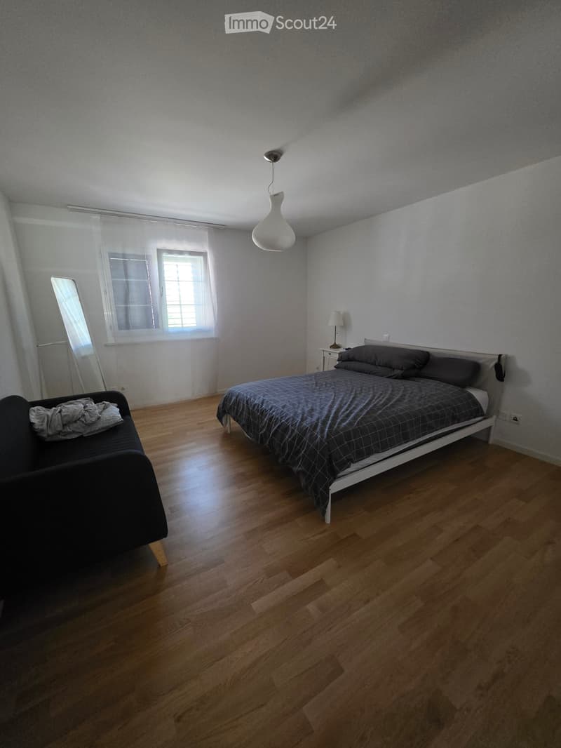 Bedroom with wooden flooring, a double bed with dark bedding, a small sofa, a nightstand with a lamp, and a mirror on the wall.