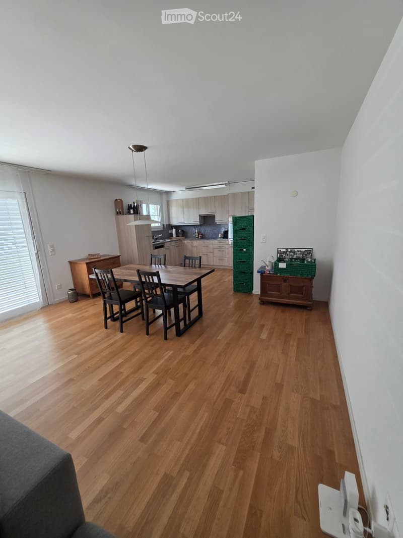 Open-plan living and dining area with wooden flooring, a dining table with chairs, kitchen cabinets, and a green crate on the floor.